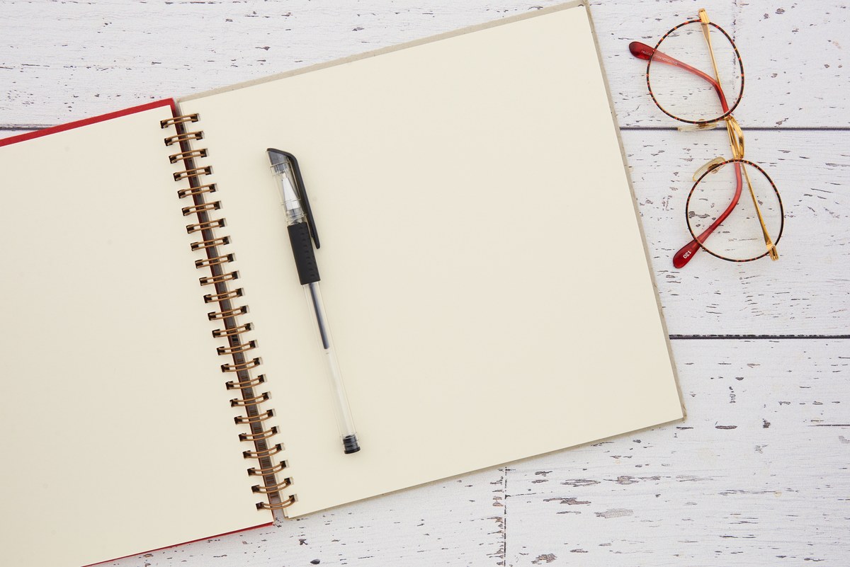 An open blank notebook with a pen and reading glasses on a pale wooden desk.
